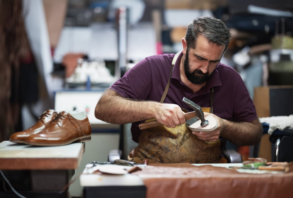 Male cobbler shaping a polished brown leather shoe at a busy workshop bench